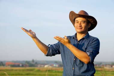 Yakışıklı Asyalı çiftçi şapka ve mavi gömlek giyer, reklam için ürünlere jest yapar. Tarım işgali kavramı. Gökyüzü arkaplanı. 