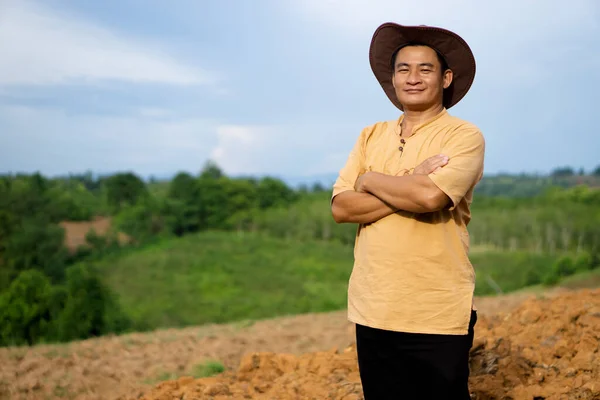 Handsome Asian man farmer is at agriculture land, wears hat and cross arms on chest, Feels confident. Concept : Agricultural occupation. Happy farmer.