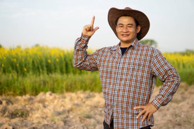Handsome Asian man farmer is at agriculture land, wears hat and plaid shirt, put hand on hip and point finger up, feels confident.  Concept : Agriculture occupation.  Copy space for adding text .