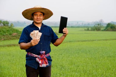 Asian man farmer is at paddy field, holds Thai banknote money and smart tablet. Concept : farmer use technology internet connection to manage crops, gets agricultural supporting money.  