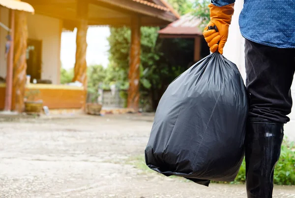 Closeup man holds black plastic bag that contains garbage inside, stand in front of house. Concept : Waste management. Environment problems. Daily chores. Throw away rubbish .