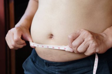 Yakından bakan adam, göbeğini ölçü bandıyla 40 inç ölçüyor. Konsept: aşırı kilolu. Obezite problemleri diğer hastalıkları etkileyebilir ya da onlara yol açabilir. Heath sorunları.         