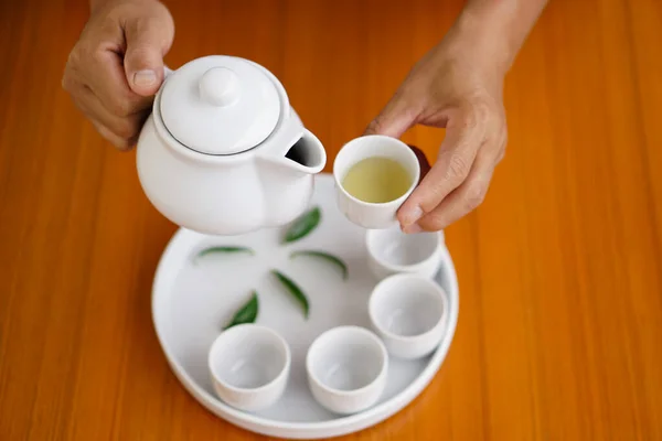Hand holds white tea pot to pour tea into cup. Wooden floor background. Concept : Tea time. Drinking tea for health. Serving traditional baverage.