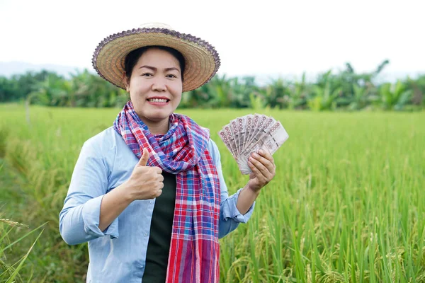 Asian farmer woman is at paddy field, hold Thai banknote money. Concept : Farmer happy to get profit, income, agriculture supporting money. Proud on crops.