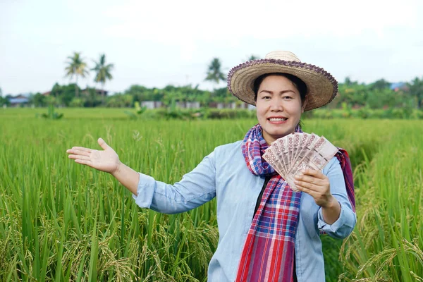 Happy Asian farmer woman is at paddy field, hold Thai banknote money. Concept : Farmer happy to get profit, income, agriculture supporting money. Proud on crops.