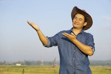 Yakışıklı Asyalı çiftçi tarım arazisinde, şapka takıyor, mavi gömlek giyiyor ve bir şey sunmak için el hareketi yapıyor. Tarım işgali kavramı. Metin veya reklam eklemek için boşluk kopyala.