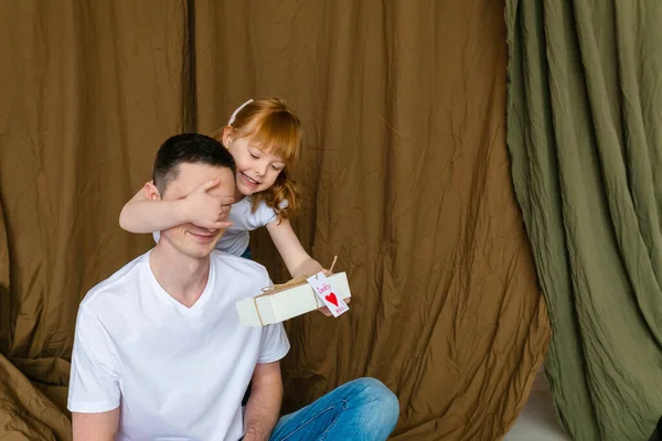 Surprise for daddy. Cute smiling caucasian child girl closes eyes of father, giving wrapped gift box and handmade postcard. Daughter congratulating dad with fathers day. Family holiday concept