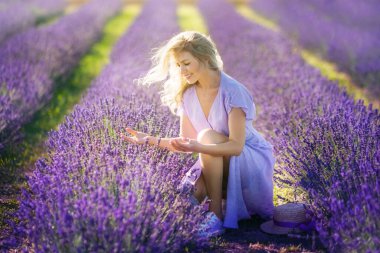 Happy young blonde woman in violet dress on blooming fragrant lavender fields with endless rows. Smiling female touching flowers. Warm sunset light. Delight in smell, aroma. Nature. Concept Happiness