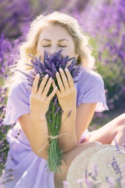 Young blonde woman holding lavender bouquet. Girl with closed eyes inhales scent of flowers. Closeup portrait. Fragrance. Romantic female in violets field, enjoying life illuminated by sunset sun rays