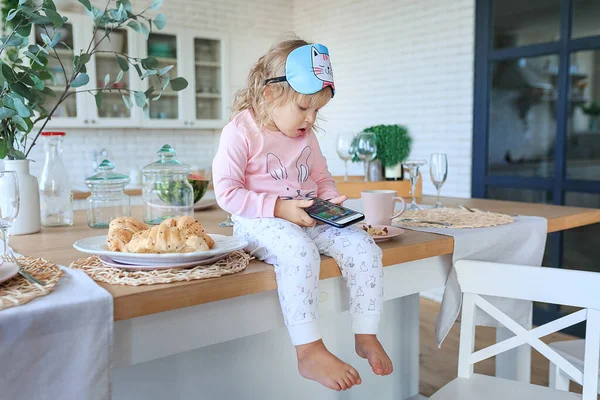 Adorable toddler eating fresh croissants and using phone. Girl dressed in pajama, sleep mask, watching cartoons while eating. Digital technologies, kids education concept. Child addiction from gadgets