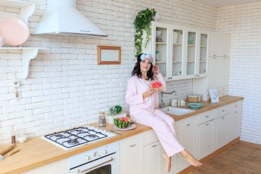 Cheerful young woman sitting on kitchen table, eating watermelon. Healthy vegetarian food, breakfast time, home wellbeing concept. Brunette female wear pajamas sleep eye mask spend weekend at home
