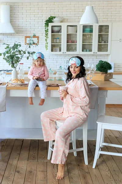 Young family in pajama, sleep mask having breakfast together. Mother, little daughter eating healthy food in morning at kitchen. Toddler kid using phone watching cartoons. Child addiction from gadgets