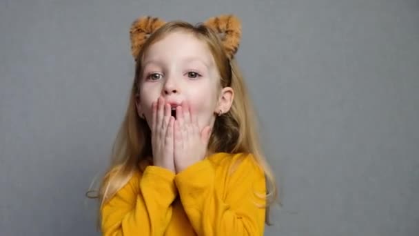 Sød overrasket blond pige med tiger ører. En hovedbeklædning med tigerører. Nytår 2022. Barn i en tiger kostume smiler sødt og ser på kameraet. – Stock-video