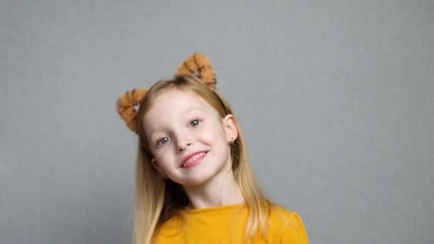 Menina loira sorridente bonito com orelhas de tigre. Uma cobertura para a cabeça com orelhas de tigre. Ano Novo 2022. Criança em um traje de tigre sorri docemente e olha para a câmera. — Vídeo de Stock
