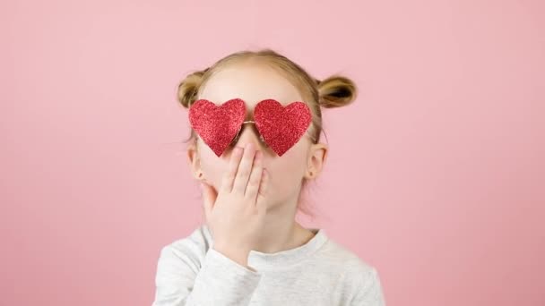 Divertente bambina bionda che invia baci aerei in occhiali da sole a forma di cuore rosso su sfondo rosa. Concetto di San Valentino o Festa della Mamma — Video Stock