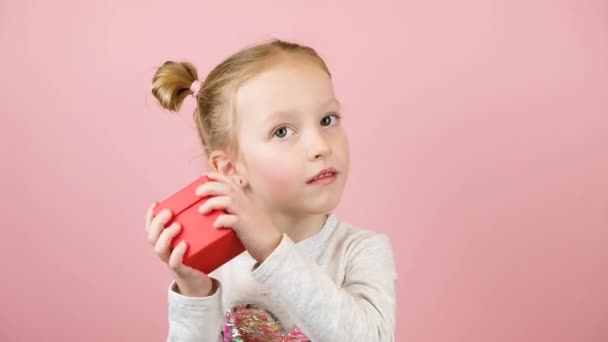 面白い好奇心旺盛な小さなブロンドの女の子は笑顔とピンクの背景に赤いギフトボックスを振る。バレンタインデーまたは母の日のコンセプト — ストック動画