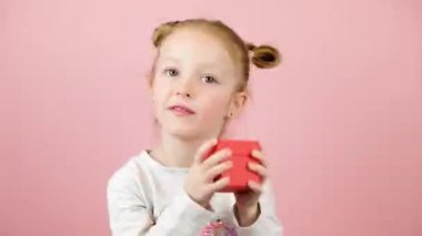 Gülen ve pembe arka planda kırmızı hediye kutusunu sallayan komik, meraklı küçük sarışın kız. Sevgililer Günü ya da Anneler Günü konsepti