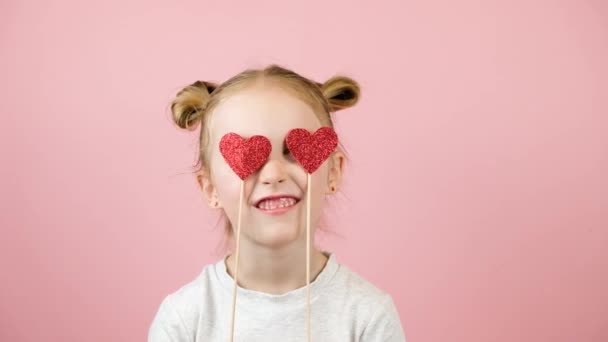핑크 배경에 빨간 하트 장난감을 가지고 웃고 놀고 있는 웃긴 금발 소녀. 발렌타인데이 또는 어머니 날의 개념 — 비디오