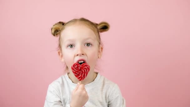 面白い小さなブロンドの女の子は笑顔とピンクの背景に赤いハート形のロリポップキャラメルを舐める。バレンタインデーまたは母の日のコンセプト — ストック動画