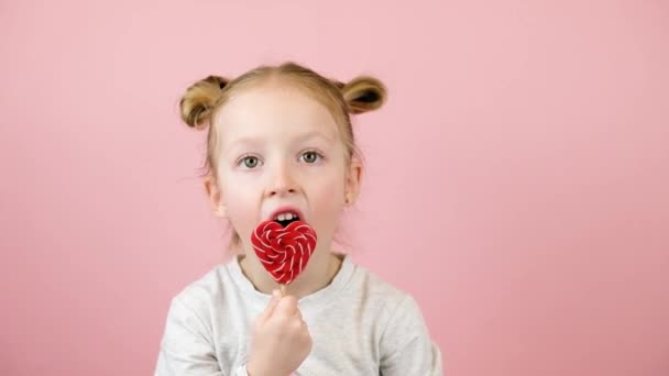 Legrační malá blondýnka s úsměvem a lízání červené srdce ve tvaru lízátka karamel na růžovém pozadí. Valentýna nebo Den matek koncept