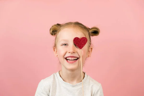Vicces kis szőke lány mosolyog, és játszik a piros szív játék rózsaszín háttér. Valentin-nap vagy anyák napja koncepció — Stock Fotó