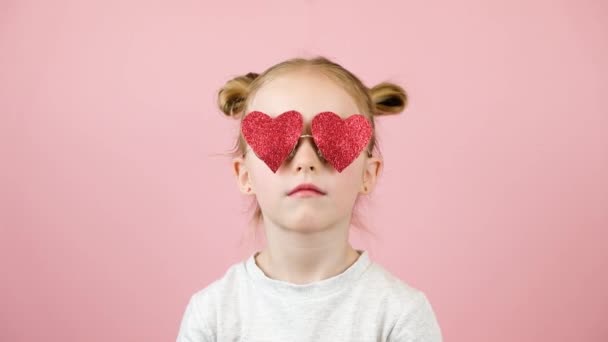 Divertida niña rubia sonriendo y jugando con gafas de sol en forma de corazón rojo sobre fondo rosa. Concepto Día de San Valentín o Día de las Madres — Vídeo de stock