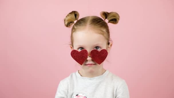 Divertida niña rubia sonriendo y jugando con gafas de sol en forma de corazón rojo sobre fondo rosa. Concepto Día de San Valentín o Día de las Madres — Vídeo de stock