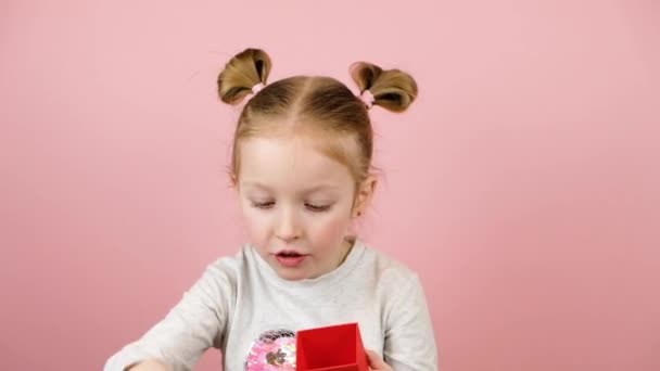 Engraçado curioso menina loira sorrindo e sacudindo a caixa de presente vermelho no fundo rosa. Conceito de Dia dos Namorados ou Dia das Mães — Vídeo de Stock