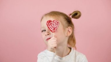 Gülen ve kırmızı kalp şekliyle oynayan komik sarışın kız pembe arka planda lolipop karamel. Sevgililer Günü ya da Anneler Günü konsepti