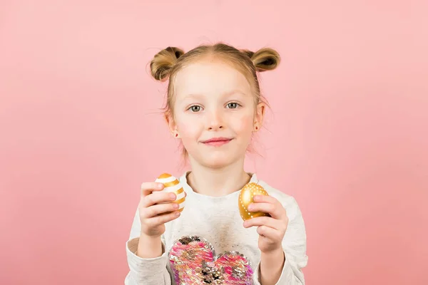 Divertida niña rubia sonriendo y jugando con huevos de Pascua dorados sobre fondo rosa. Feliz concepto de Pascua — Foto de Stock