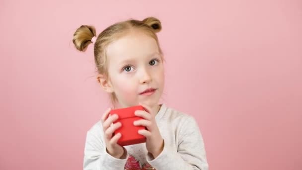 面白い好奇心旺盛な小さなブロンドの女の子は笑顔とピンクの背景に赤いギフトボックスを振る。バレンタインデーまたは母の日のコンセプト — ストック動画