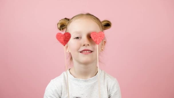 Menina loira engraçada sorrindo e brincando com o brinquedo do coração vermelho no fundo rosa. Conceito de Dia dos Namorados ou Dia das Mães — Vídeo de Stock