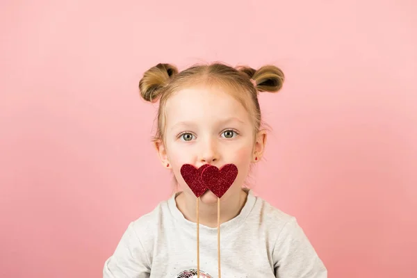 Vicces kis szőke lány mosolyog, és játszik a piros szív játék rózsaszín háttér. Valentin-nap vagy anyák napja koncepció — Stock Fotó