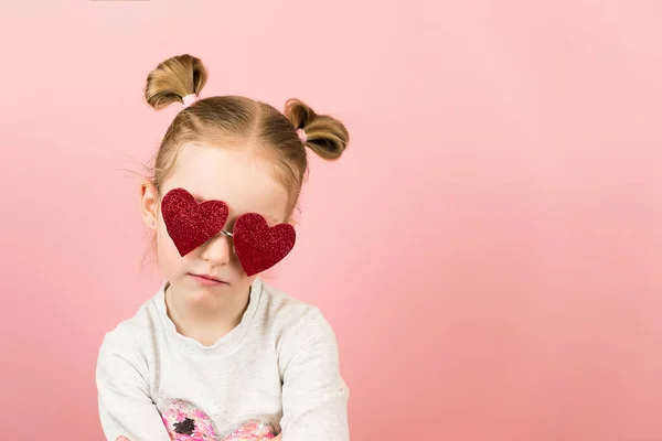 Vicces, dühös szőke lány piros szív alakú napszemüvegben, rózsaszín háttérrel. Valentin-nap vagy anyák napja koncepció — Stock Fotó