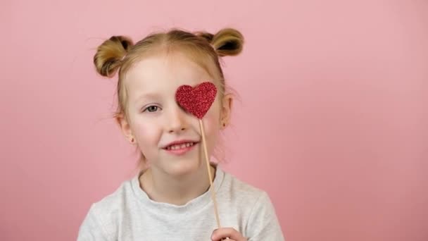 面白い小さなブロンドの女の子は笑顔とピンクの背景に赤いハートのおもちゃで遊んでいます。バレンタインデーまたは母の日のコンセプト — ストック動画