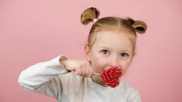 Gülen ve kırmızı kalp şekliyle oynayan komik sarışın kız pembe arka planda lolipop karamel. Sevgililer Günü ya da Anneler Günü konsepti — Stok video