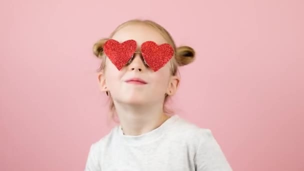 Kırmızı kalp şeklinde gözlüklerle pembe arka planda gülümseyen ve dans eden komik sarışın kız. Sevgililer Günü ya da Anneler Günü konsepti — Stok video
