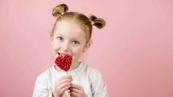 Petite fille blonde drôle souriante et léchant caramel sucette en forme de coeur rouge sur fond rose. Concept de Saint Valentin ou Fête des Mères — Video