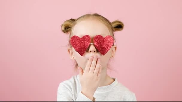 Funny little blonde girl sending air kiss in red heart shape sunglasses on pink background. Valentines Day or Mothers Day concept — Stock Video