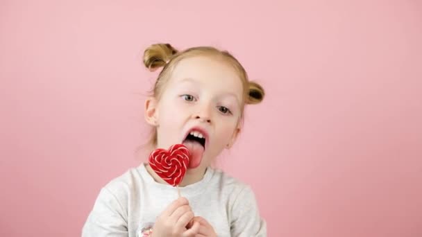 Chica rubia divertida sonriendo y lamiendo caramelo lollipop en forma de corazón rojo sobre fondo rosa. Concepto Día de San Valentín o Día de las Madres — Vídeo de stock