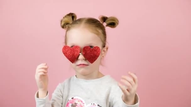 Rolig liten blond flicka ler och leker med röda hjärta form solglasögon på rosa bakgrund. Alla hjärtans dag eller mors dag koncept — Stockvideo