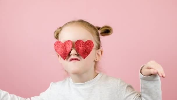 Дивна дівчинка-блондинка посміхається і танцює в сонцезахисних окулярах червоної серцевої форми на рожевому тлі. День святого Валентина або День матері — стокове відео
