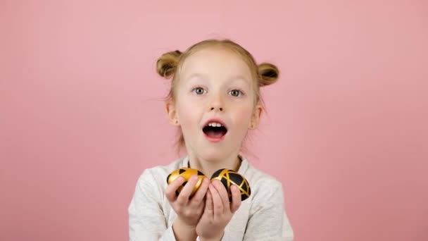 面白い小さなブロンドの女の子は笑顔で、ピンクの背景に黄金のイースターエッグで遊んでいます。ハッピーイースターのコンセプト — ストック動画