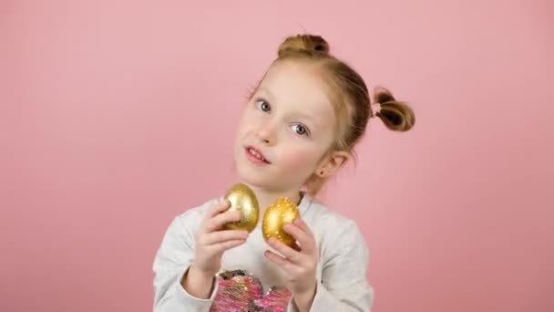Αστείο μικρό ξανθό κορίτσι χαμογελά και παίζει με χρυσά αυγά Πάσχα σε ροζ φόντο. Καλή ιδέα Πάσχα — Αρχείο Βίντεο