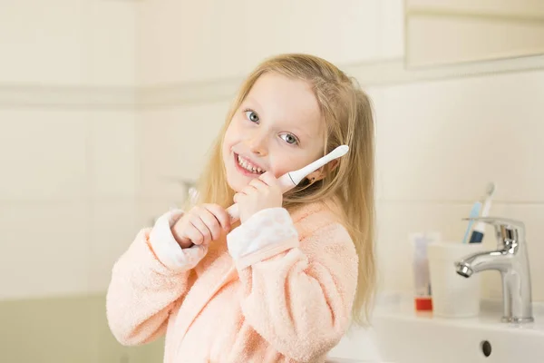 Ευτυχισμένο χαμογελαστό κοριτσάκι βουρτσίζουν τα δόντια χρησιμοποιώντας υπερήχων ηλεκτρική οδοντόβουρτσα στο μπάνιο στο σπίτι. Στοματική υγιεινή, υγεία δοντιών και ούλων, υγιή δόντια. Καθημερινή ζωή και ρουτίνα. — Φωτογραφία Αρχείου