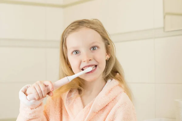 Szczęśliwie uśmiechnięta dziewczynka myje zęby używając ultradźwiękowej elektrycznej szczoteczki do zębów w łazience w domu. Higiena jamy ustnej, zdrowie zębów i dziąseł, zdrowe zęby. Codzienne życie i rutyna. — Zdjęcie stockowe