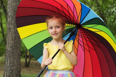 Happy funny girl holding rainbow umbrella. Cute Happy schoolgirl playing in rainy summer park. Kid walking in autumn shower. Outdoor fun by any weather.