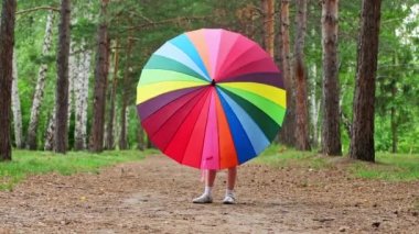 Happy funny girl holding rainbow umbrella. Cute Happy schoolgirl playing in rainy summer park. Kid walking in autumn shower. Outdoor fun by any weather.