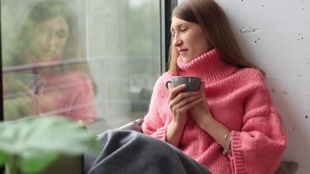 雨の日は窓からお茶を飲んでいる女の子 秋の気分 暖かいセーターと居心地の良い雰囲気 窓の近くに座っている — ストック動画