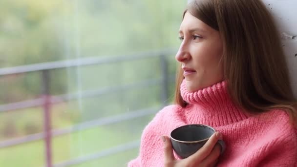 雨の日は窓からお茶を飲んでいる女の子 秋の気分 暖かいセーターと居心地の良い雰囲気 少女の肖像画 — ストック動画